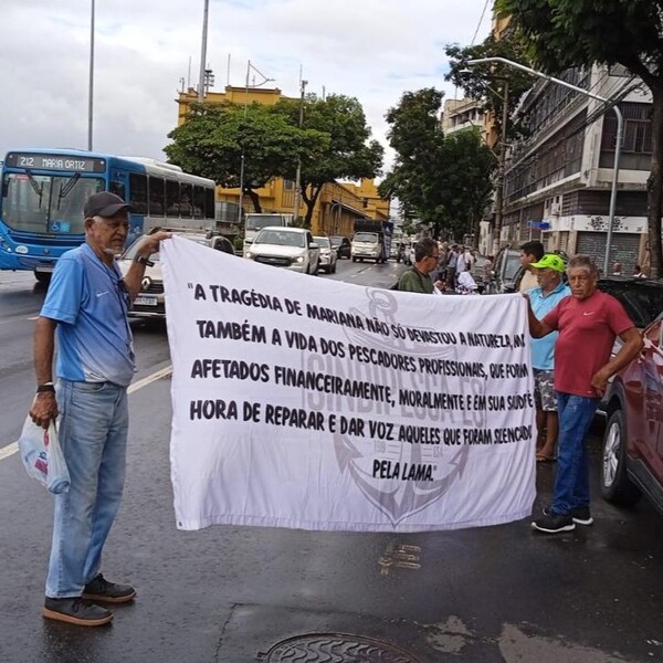 Dois homens seguram uma faixa branca com texto sobre a tragédia de Mariana e seus impactos nos pescadores profissionais, em uma rua movimentada com veículos ao fundo.