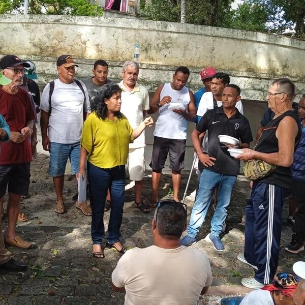Grupo de pessoas em uma área externa, algumas em pé e outras sentadas, em um ambiente com árvores e paredes antigas.