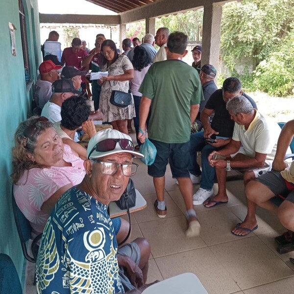 Pessoas sentadas e em pé em um corredor, algumas conversando e outras observando, em um ambiente coberto com paredes verdes.