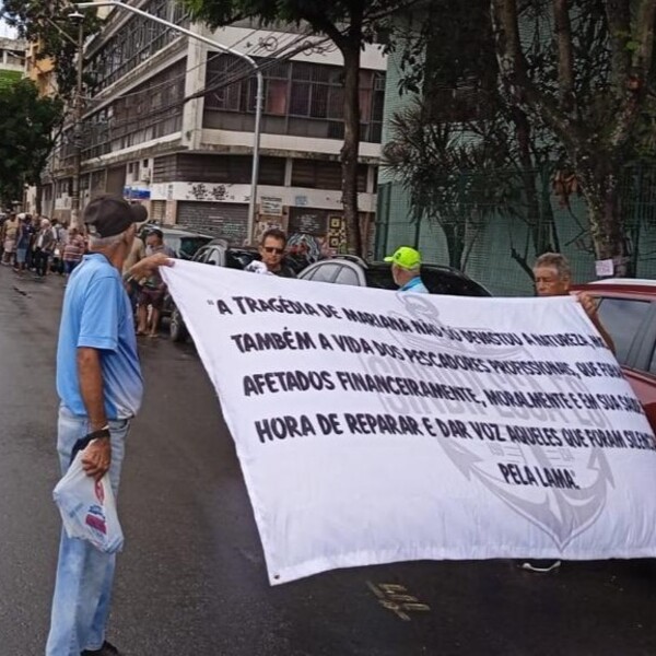Dois homens seguram uma faixa branca com texto sobre a tragédia de Mariana, em uma rua com edifícios antigos e árvores ao fundo.
