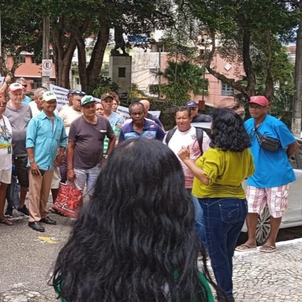 Grupo de pessoas reunidas ao ar livre, algumas ouvindo uma mulher de camisa verde, com árvores e prédios ao fundo.